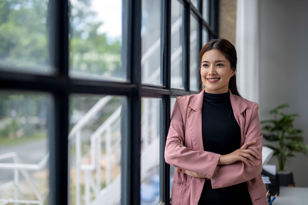 Une femme en veste rose se tient devant une fenêtre, les bras croisés.